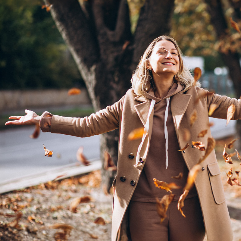 Donna sorridente con le braccia aperte mentre gioca con le foglie autunnali, simbolo di energia ritrovata e benessere.