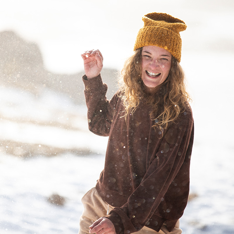 Giovane donna sorridente gioca nella neve con cappello giallo, rappresentando vitalità e benessere invernale.