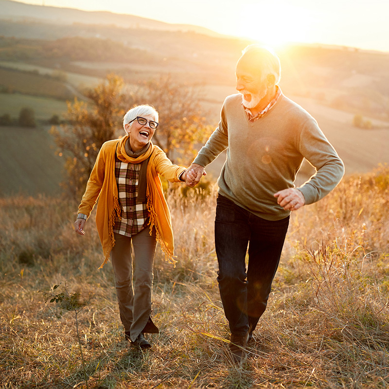 Due persone anziane sorridenti camminano all’aperto in un parco d’autunno, in simbolo di energia e vitalità.